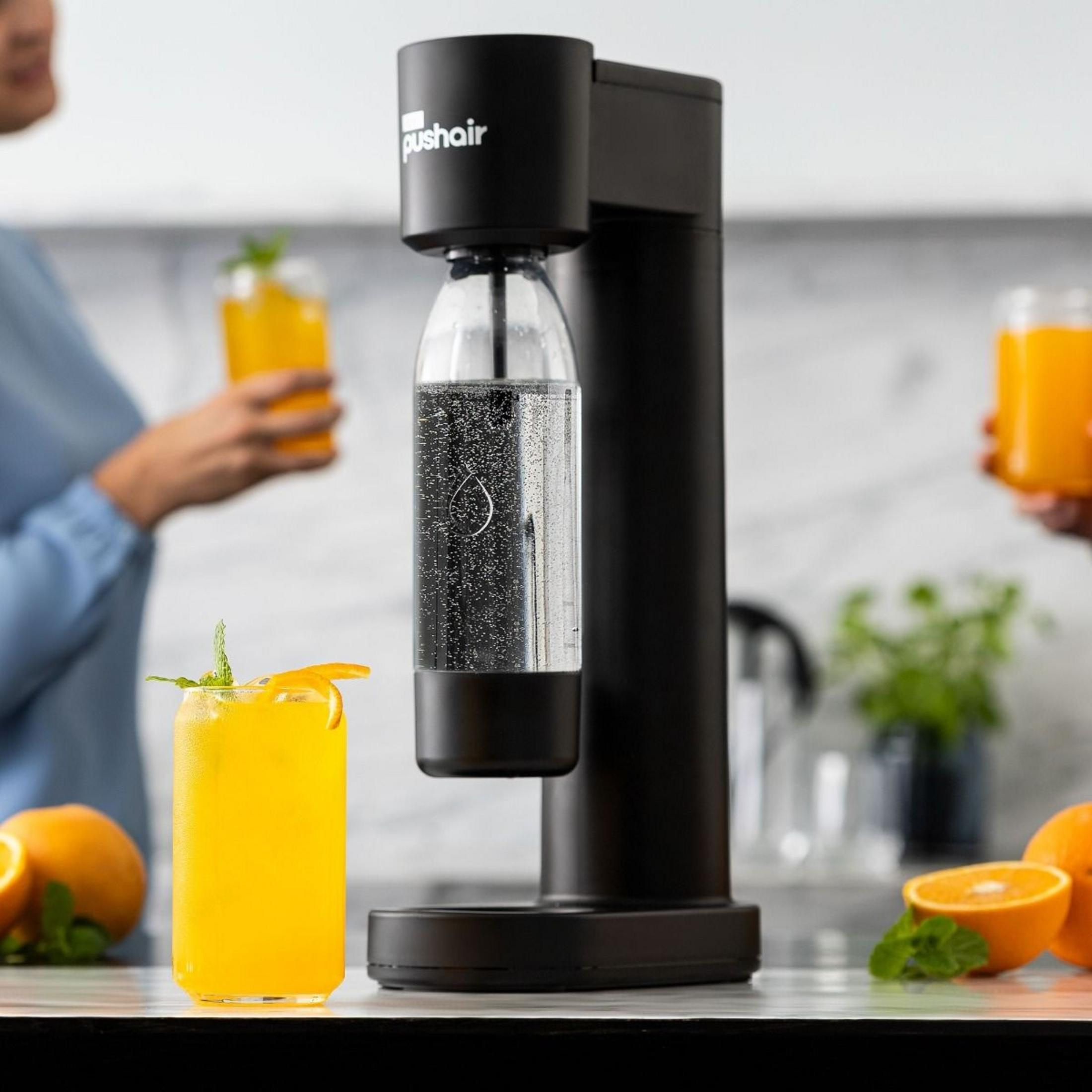 Ein schwarzer Wassersprudler steht auf einer Theke, flankiert von Gläsern mit Orangensaft und frischen Orangen.
