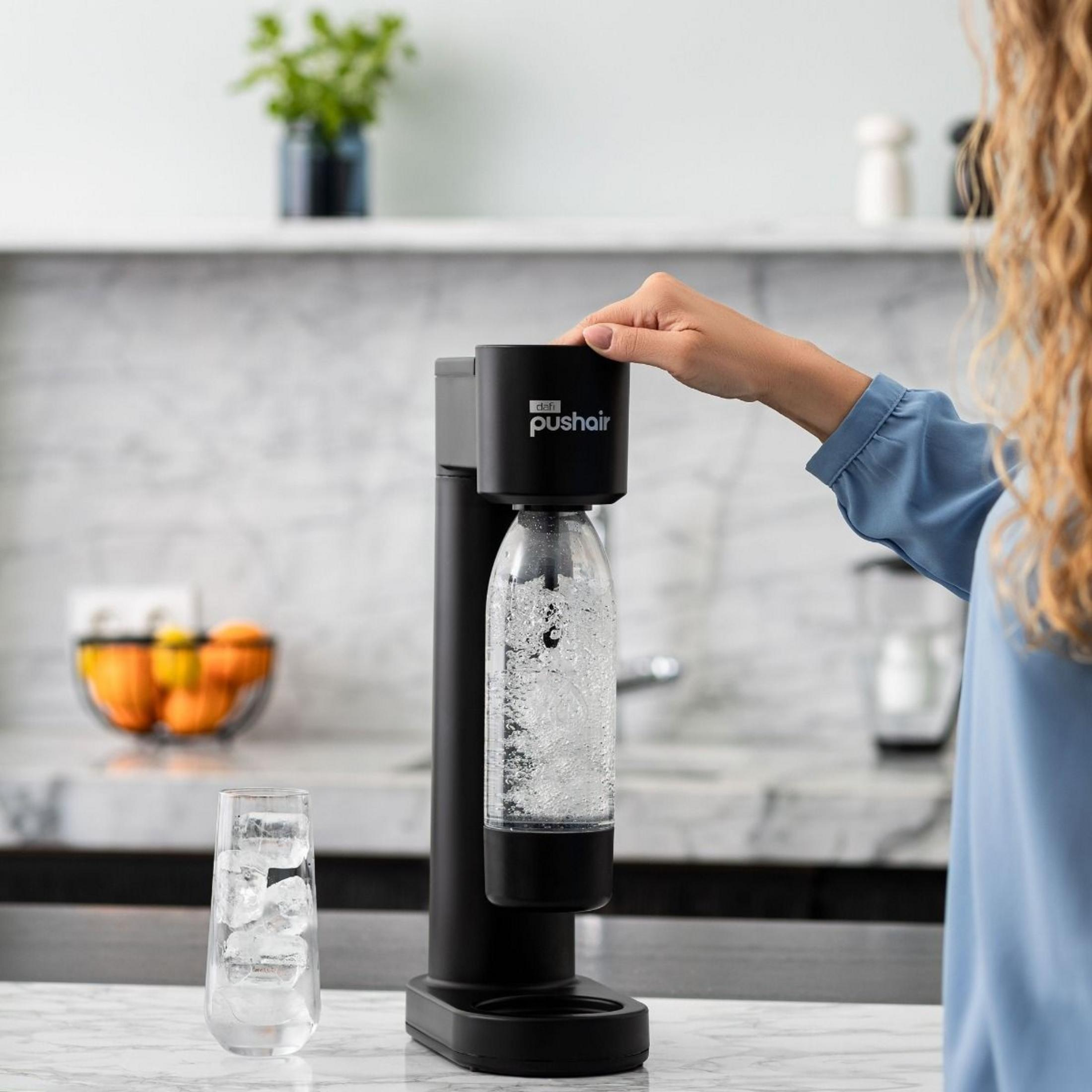 Eine Frau drückt den Knopf eines schwarzen Wassersprudlers auf einer weißen Küchentheke mit einem Eisglas daneben.