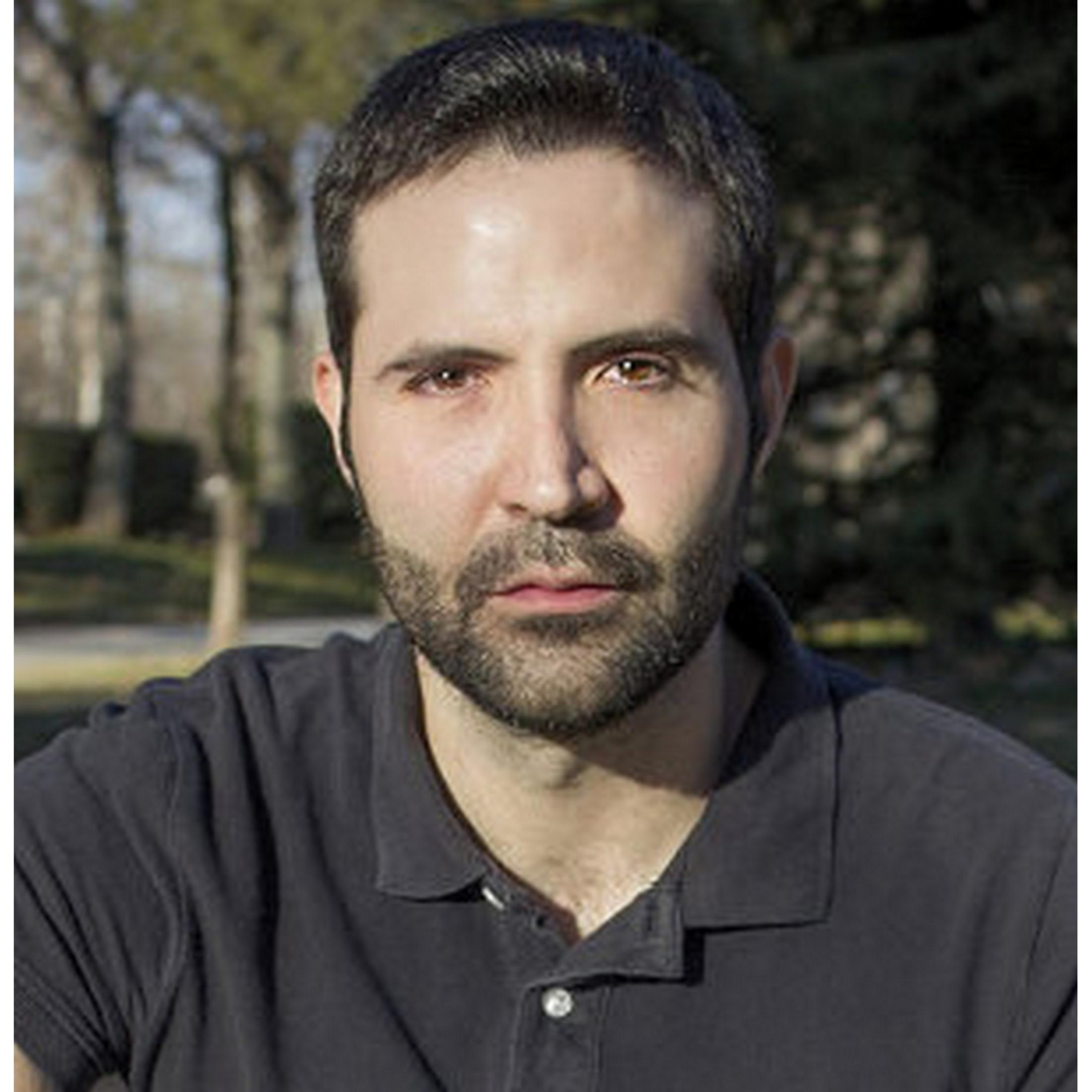 Primer plano de un hombre con pelo oscuro y barba corta, con polo oscuro, al aire libre con fondo difuminado de árboles.