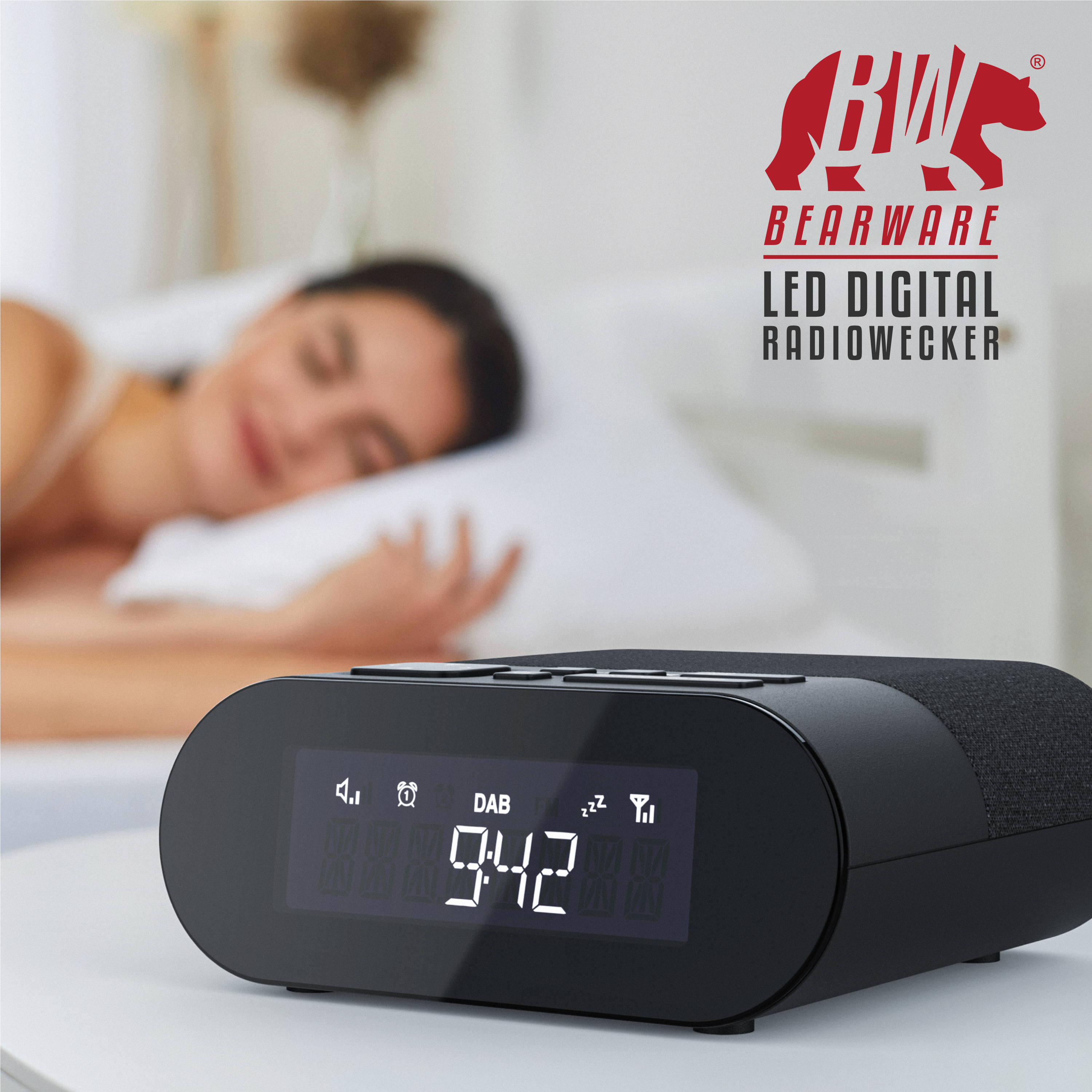 Ein digitaler Radiowecker auf einem Tisch, im Hintergrund schläft eine Frau im Bett.
