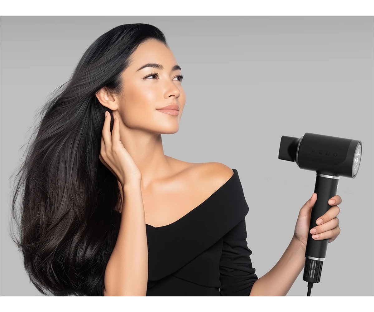 Kobieta z długimi ciemnymi włosami trzyma czarną suszarkę do włosów na szarym tle.