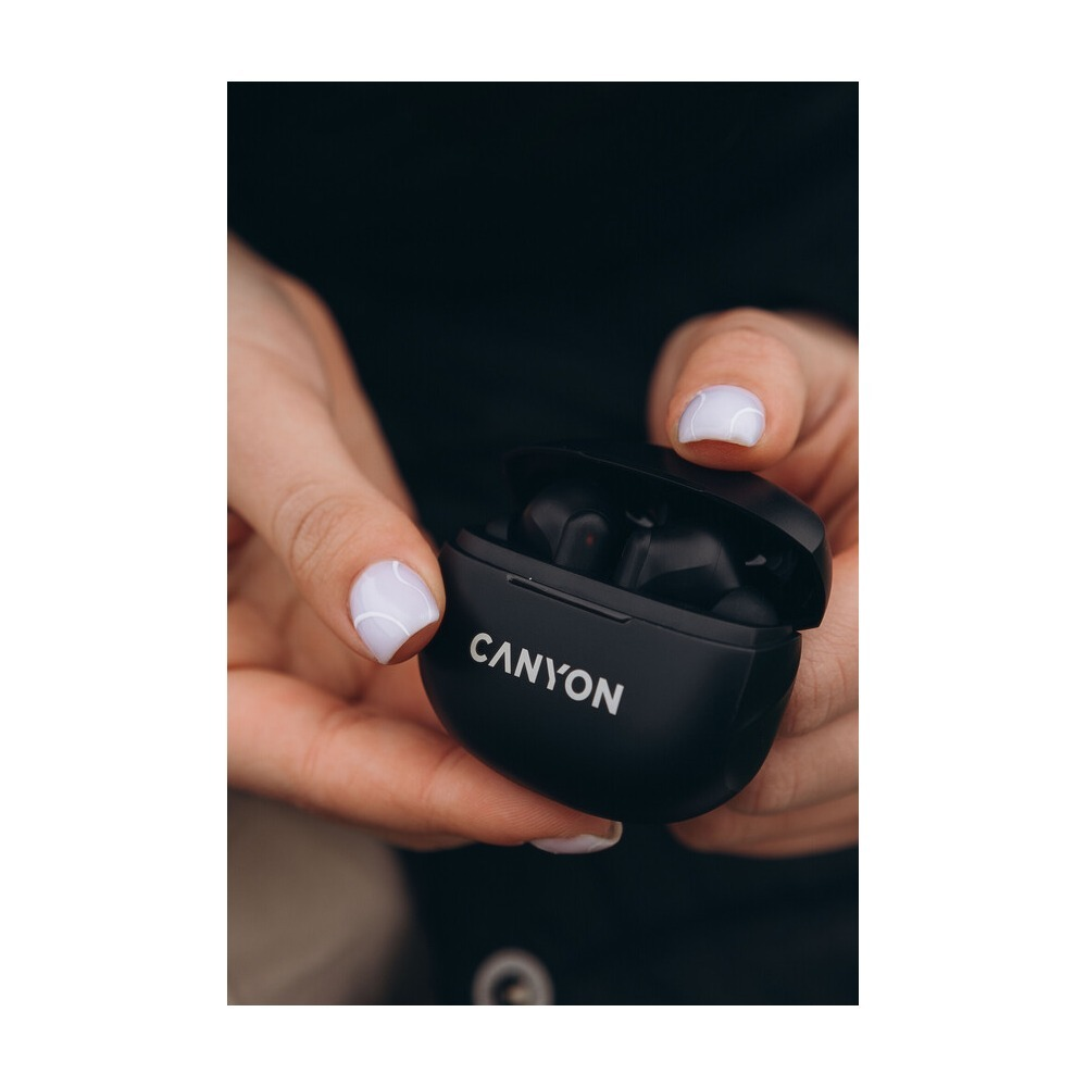 Ręce trzymające otwarte czarne etui ładujące do słuchawek bezprzewodowych Canyon z białym logo.