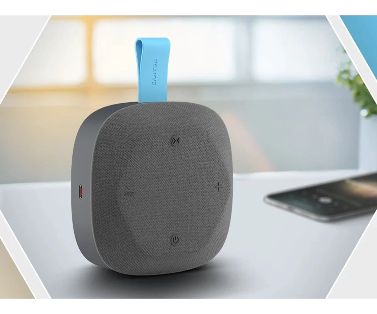 Szary, kwadratowy głośnik Bluetooth z niebieskim paskiem do noszenia na białym stole.