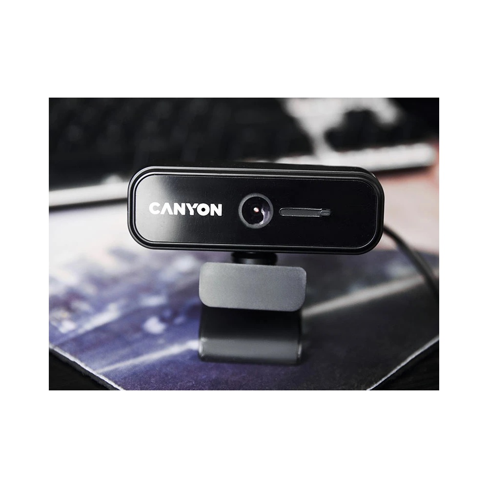Czarna kamera internetowa Canyon z wbudowaną podstawką, na rozmytym tle biurka i klawiatury.
