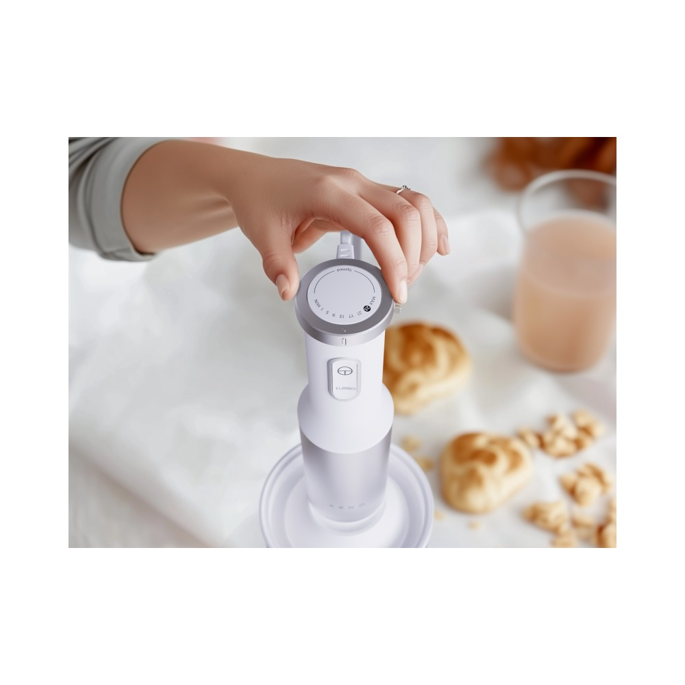 Dłoń trzyma biały blender ręczny z regulacją prędkości na białym stole obok wypieków i szklanki napoju.