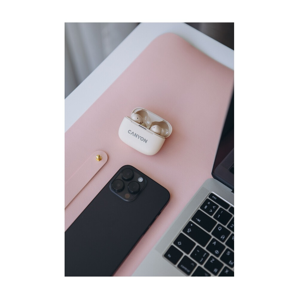 Beżowe słuchawki Canyon w etui, czarny smartfon i laptop leżą na różowej podkładce na biurko.