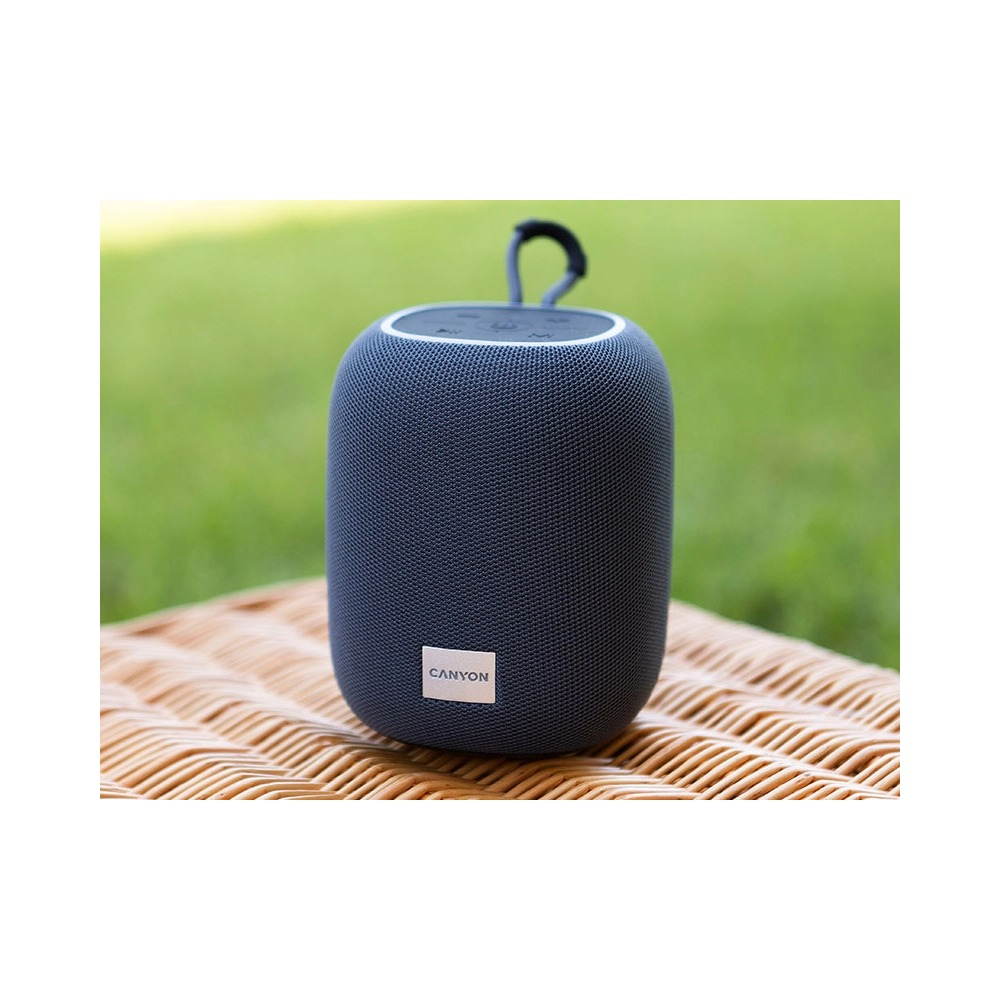Szary głośnik Bluetooth Canyon z paskiem do noszenia na wiklinowym koszu na zielonym tle.