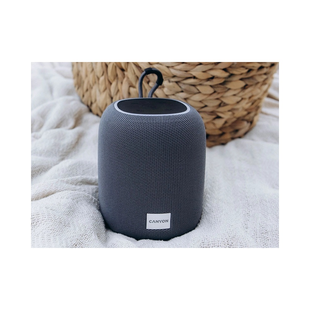 Szary głośnik bluetooth Canyon na białym kocu, w tle pleciony koszyk.