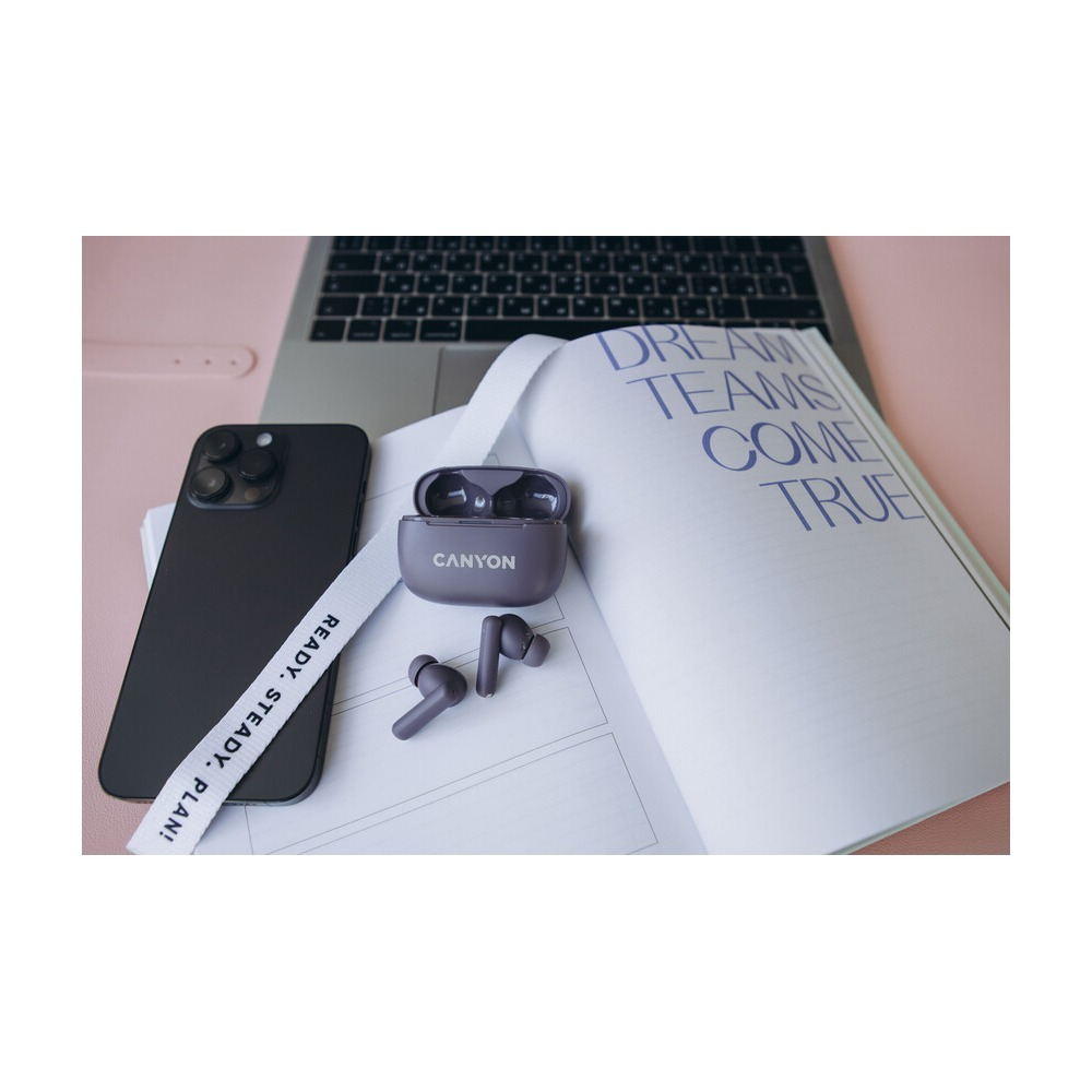 Fioletowe słuchawki Canyon w etui ładującym na książce obok czarnego smartfona i laptopa.