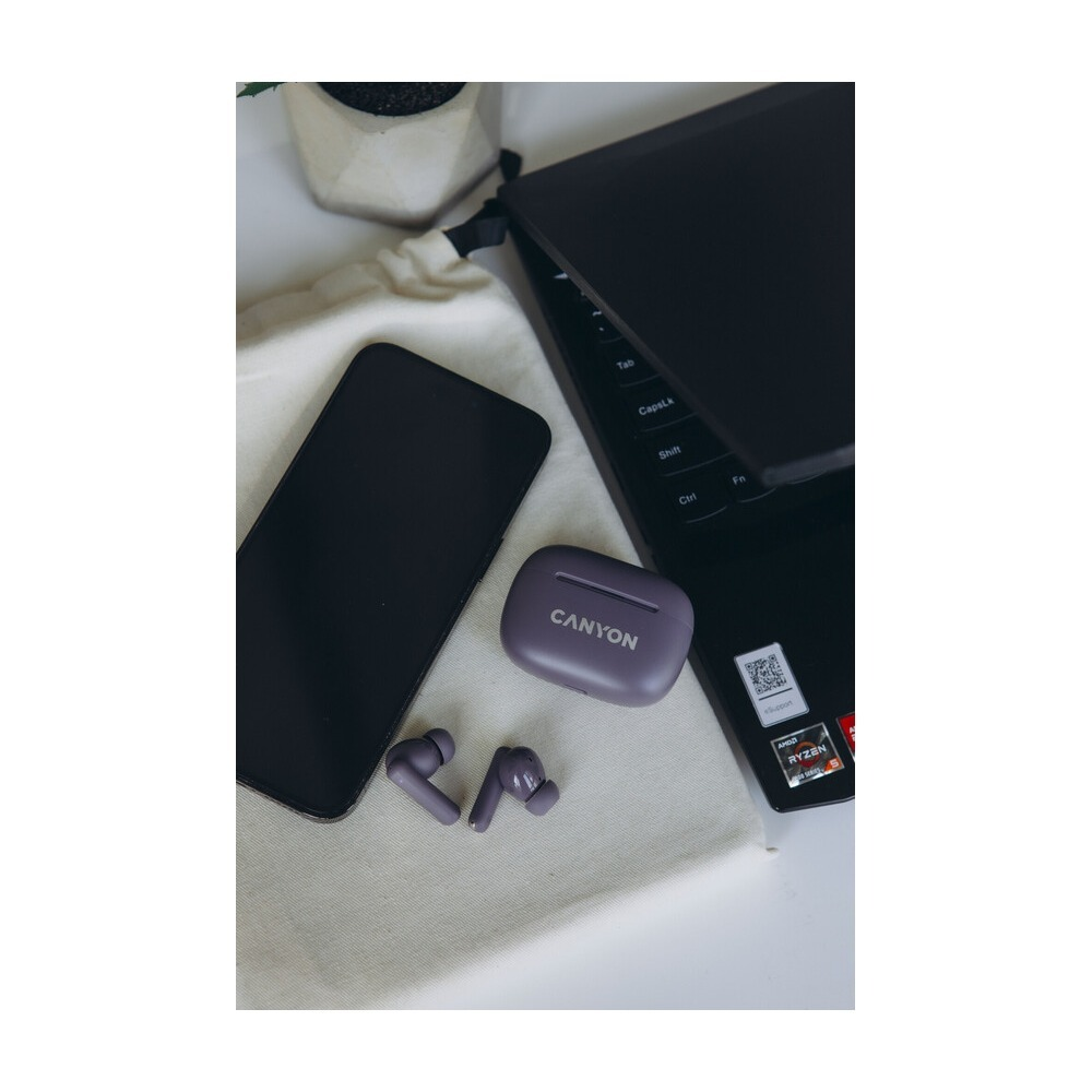 Czarny smartfon, fioletowe słuchawki Canyon z etui i laptop leżą na materiałowej torbie.