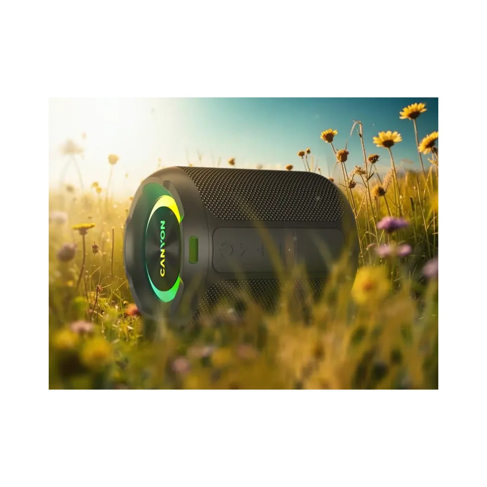 Cylindryczny głośnik Bluetooth Canyon z zielonym światłem, umieszczony na słonecznym polu kwiatów.