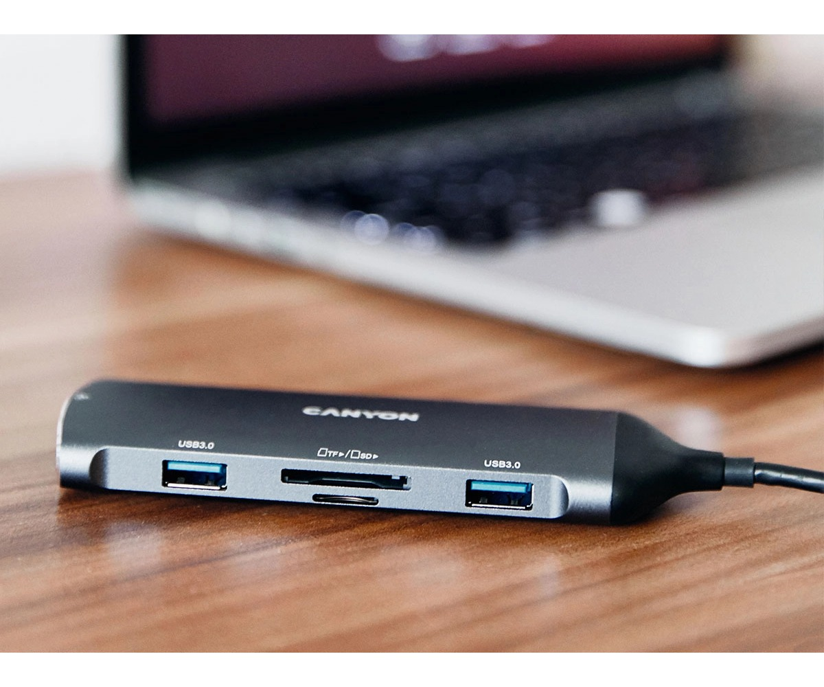 Szary hub USB Canyon z dwoma portami USB 3.0 i czytnikiem kart na drewnianym biurku przed laptopem.