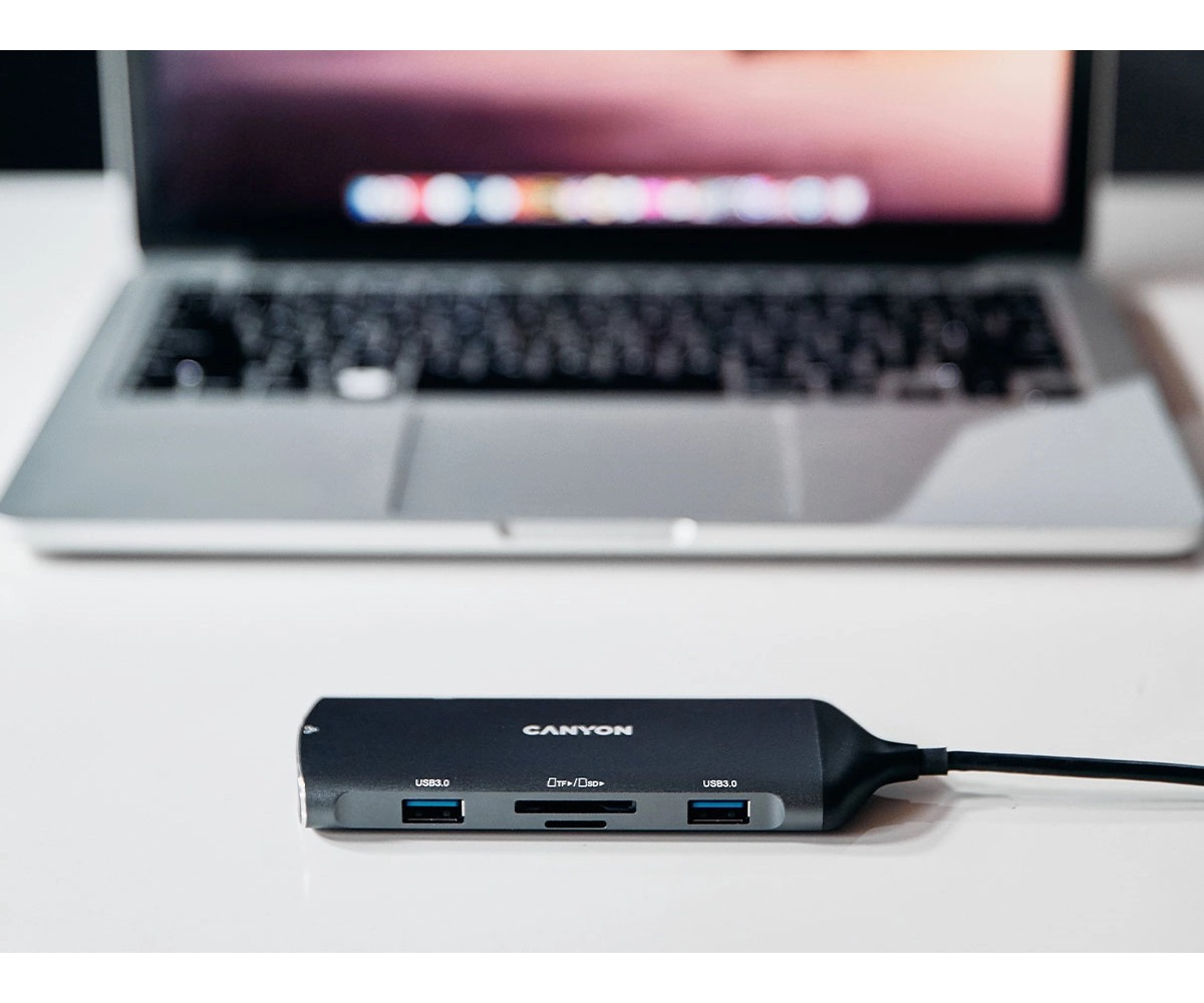 Ciemny hub USB Canyon z portami USB i czytnikiem kart leży na białym stole przed laptopem.