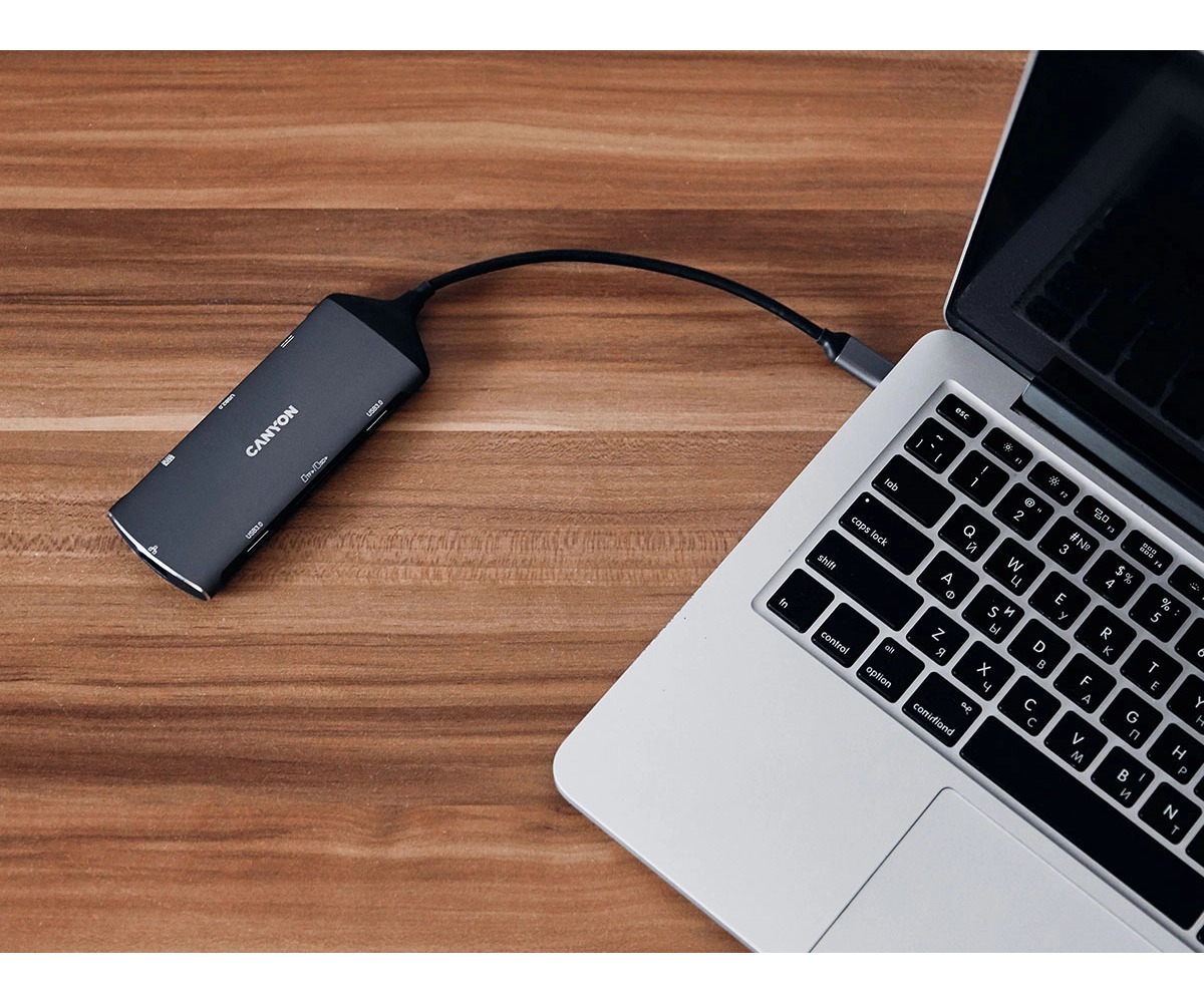 Hub USB-C Canyon leży na drewnianym biurku i jest podłączony do laptopa.