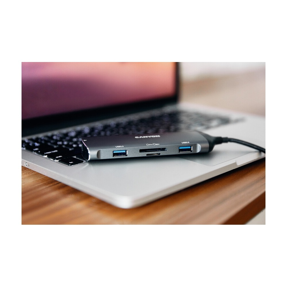 Szary hub USB-C spoczywa na klawiaturze srebrnego laptopa umieszczonego na drewnianym stole.