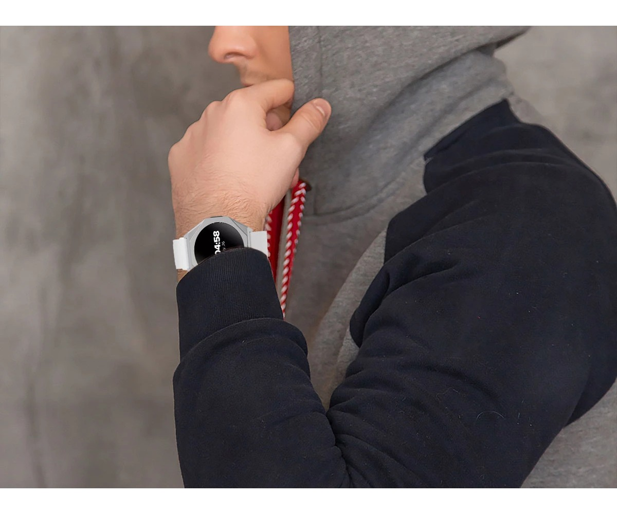 Osoba w szarej bluzie z kapturem i niebieskim swetrze nosi biały smartwatch na nadgarstku na tle szarej ściany.