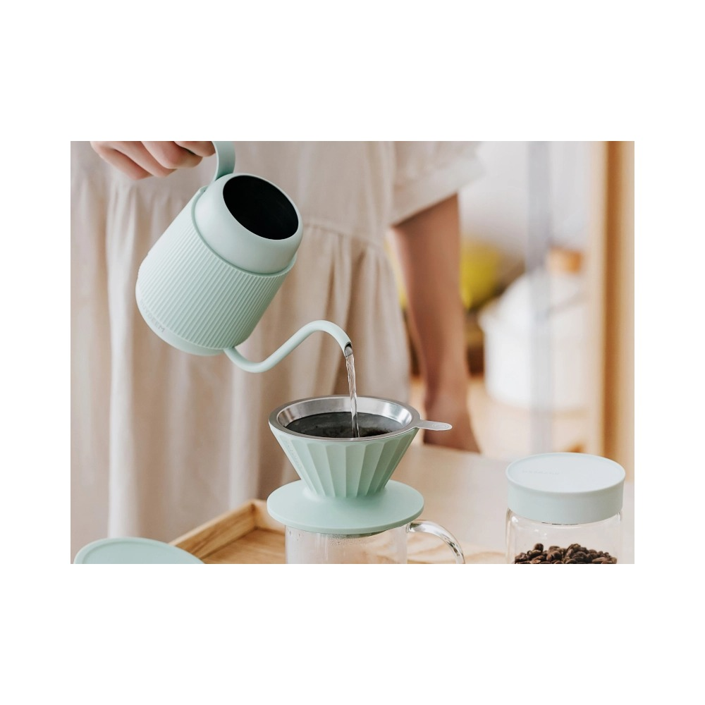 Una persona versa acqua da un bollitore verde chiaro in un filtro per caffè coordinato su un vassoio di legno.