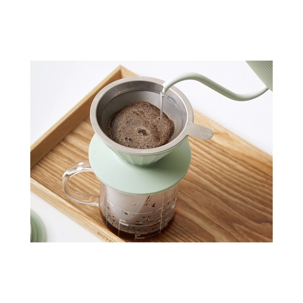 L'acqua viene versata da un bollitore verde chiaro in un colatoio per caffè su un vassoio di legno.