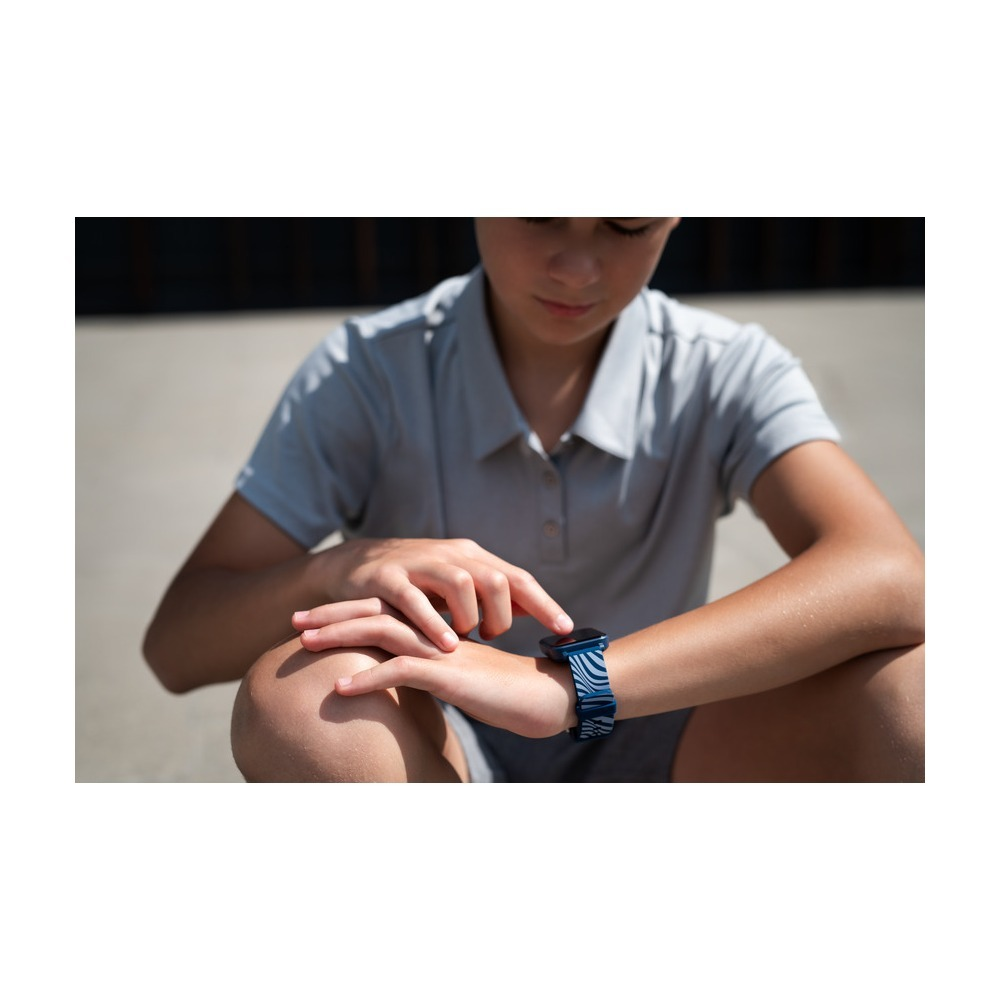 A boy in a gray shirt sits outdoors, tapping on a blue-and-white patterned smartwatch display.