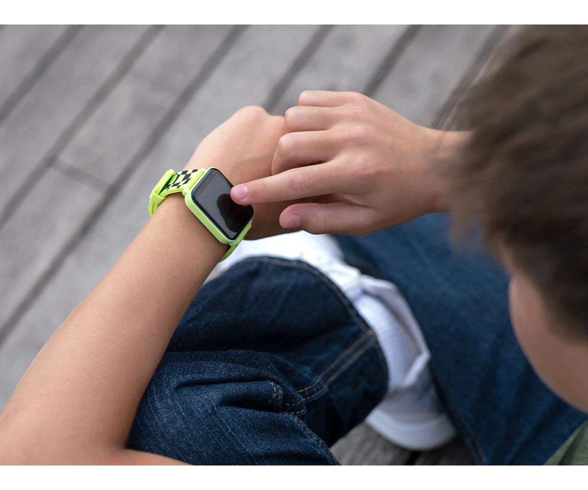 A child taps a neon green smartwatch on their wrist while sitting on wooden planks.