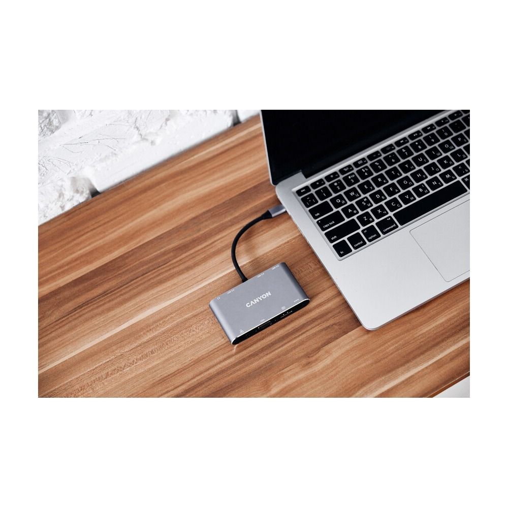 Srebrny hub USB Canyon jest podłączony do laptopa na drewnianej powierzchni.