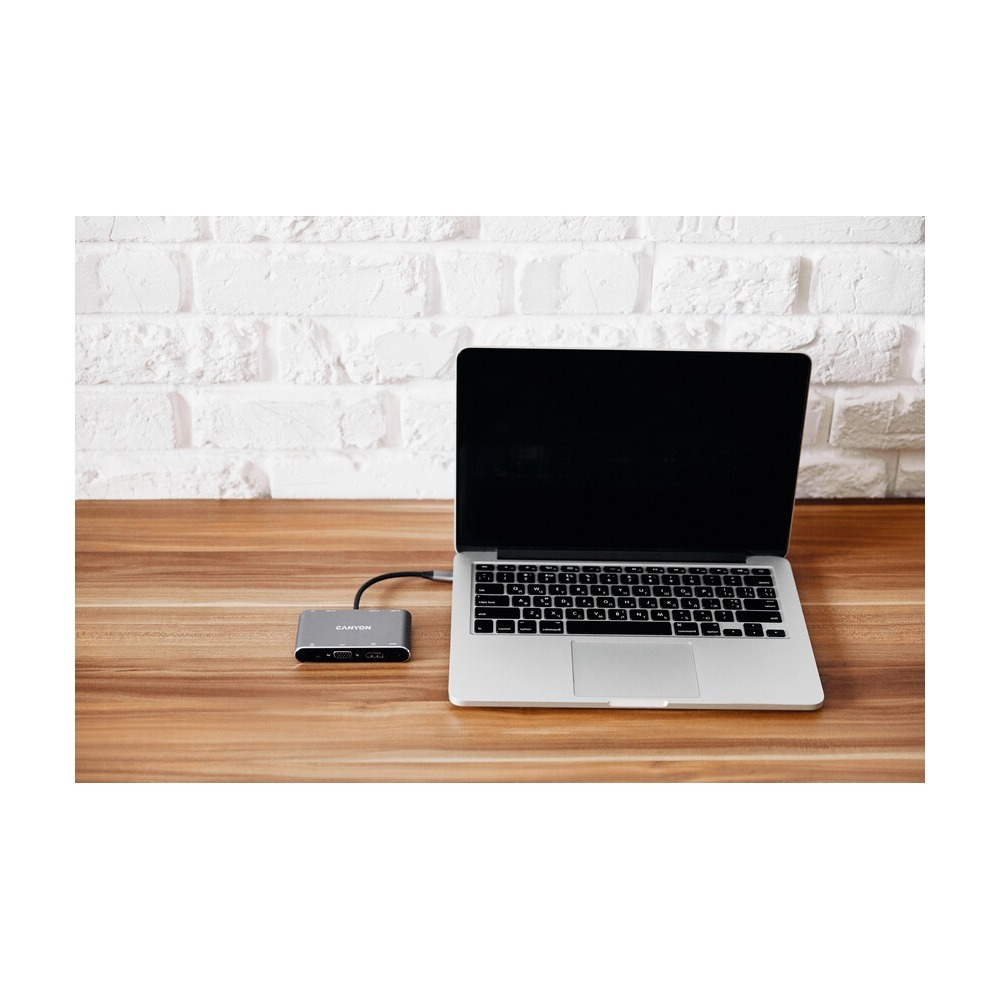 Srebrny laptop i hub USB-C Canyon stoją na drewnianym biurku na tle białej ściany z cegły.