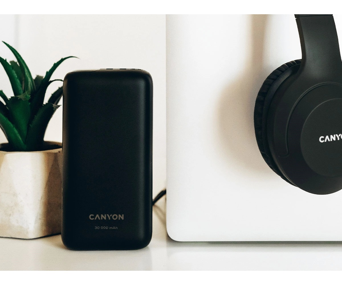 Czarny powerbank Canyon stoi obok rośliny w doniczce i czarnych słuchawek.