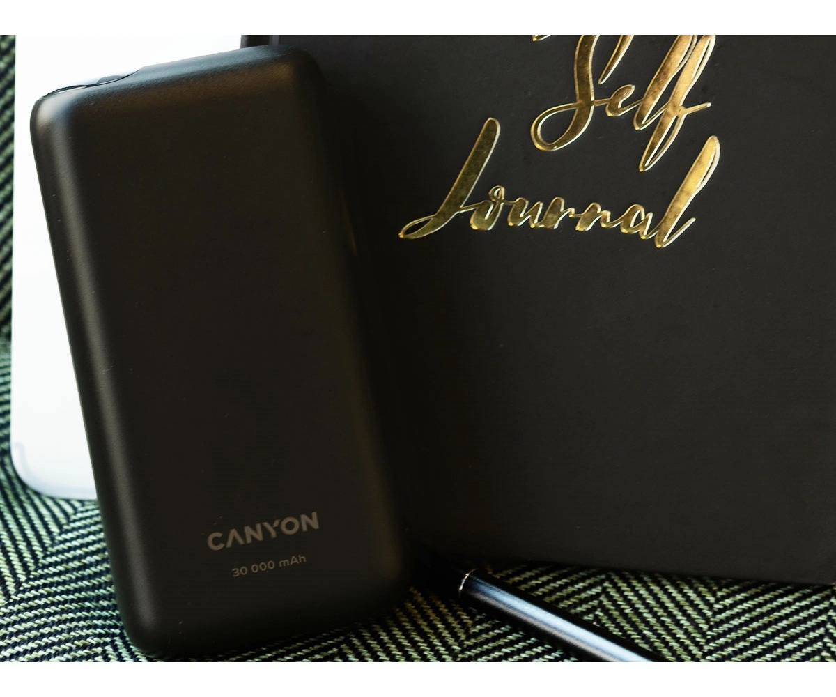 Czarny powerbank Canyon 30 000 mAh stoi obok czarnego dziennika z złotym napisem i długopisem na zielonej powierzchni.