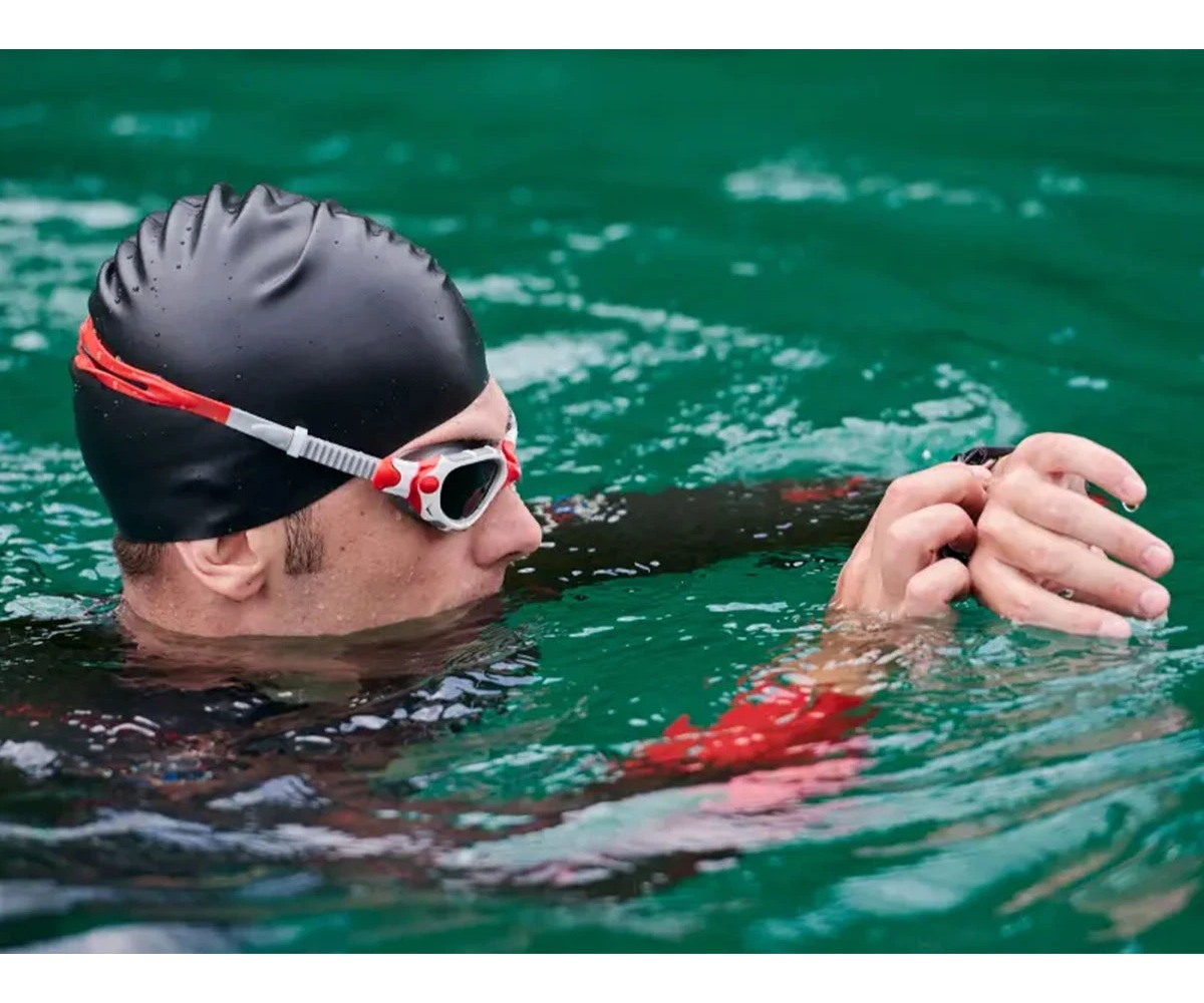 Un nadador con gorro de baño negro y gafas en agua verde revisa su reloj.