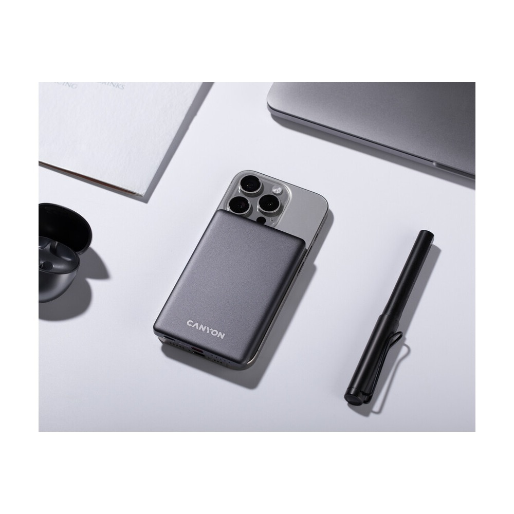 Szary power bank Canyon przymocowany do smartfona na białym biurku z długopisem i słuchawkami.