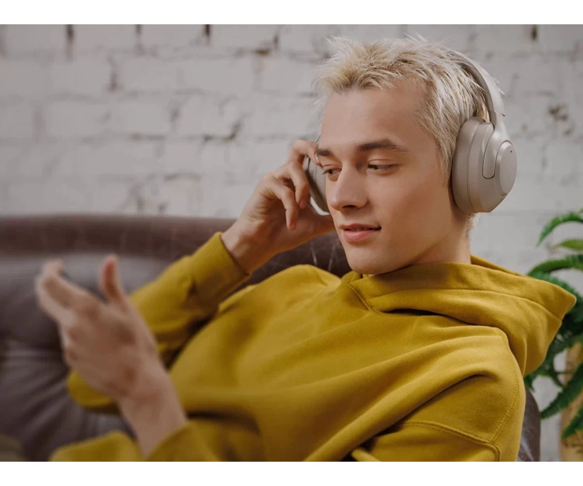 Blondyn w żółtej bluzie z kapturem nosi duże białe słuchawki i patrzy na smartfon.