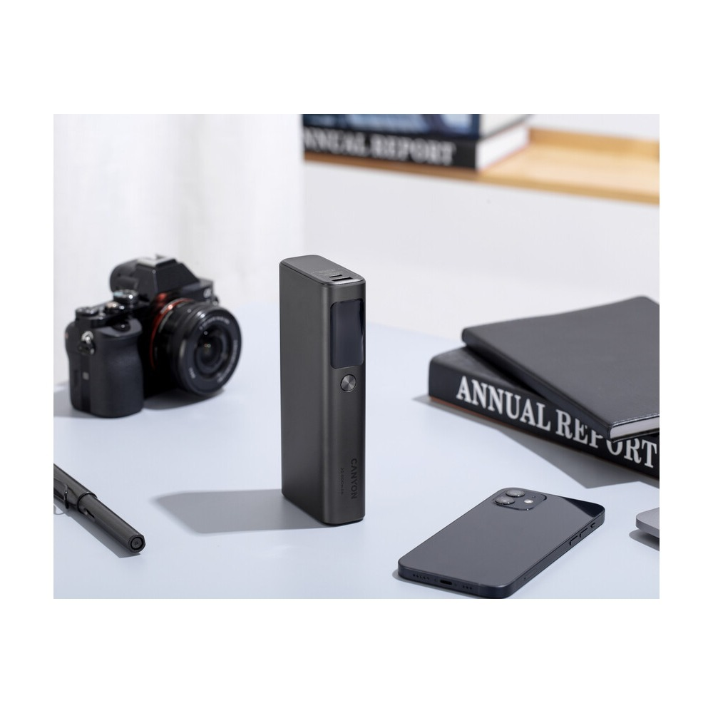 Szary power bank stoi na biurku, otoczony aparatem, smartfonem, piórem i książkami.