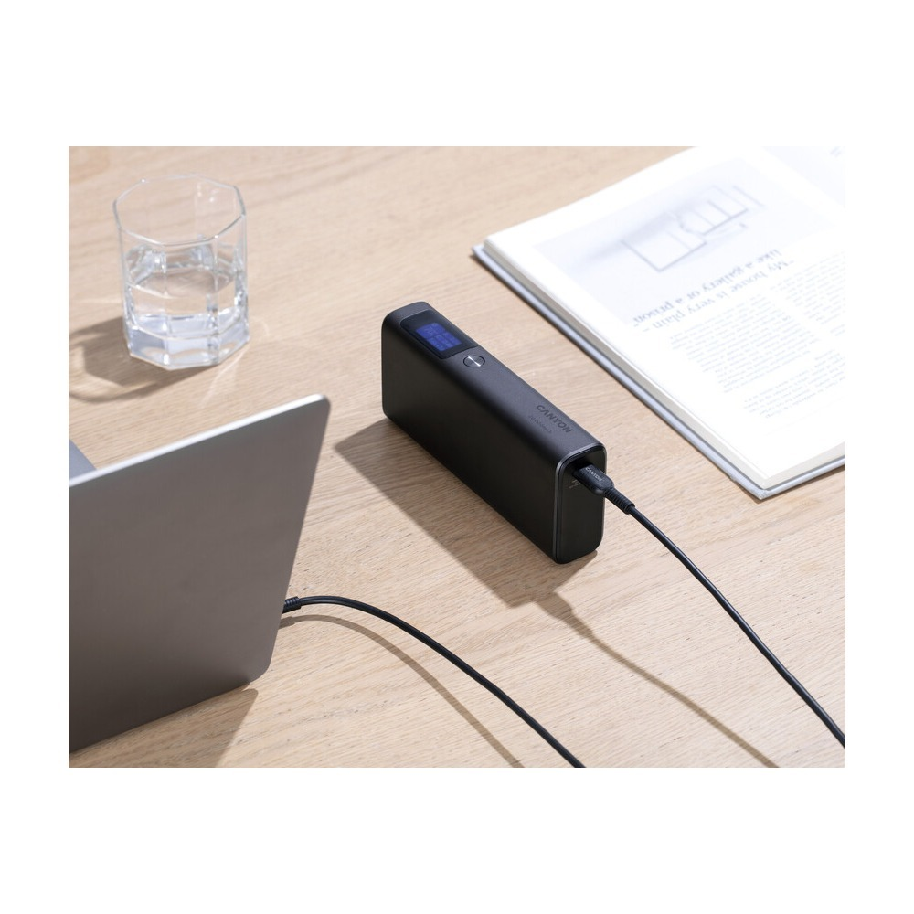 Czarny power bank na drewnianym stole, połączony kablem, obok szklanki i otwartej książki.