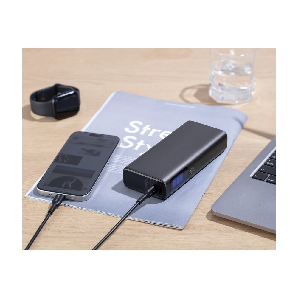 Powerbank na czasopiśmie ładuje smartfon na drewnianym stole z laptopem i zegarkiem.