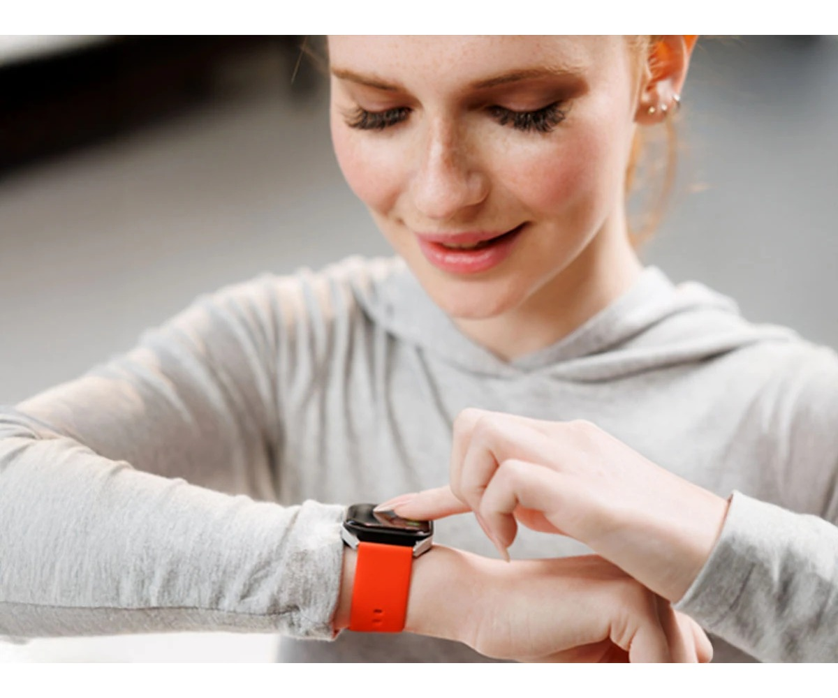 Uśmiechnięta kobieta w szarej bluzie patrzy na smartwatch z pomarańczowym paskiem na nadgarstku.