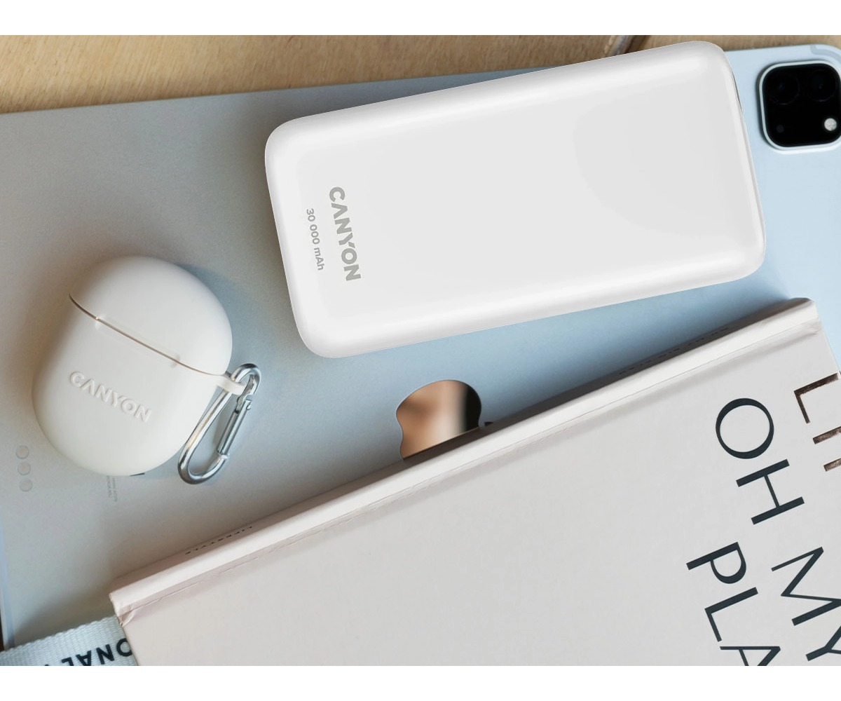 Biały powerbank Canyon i etui ładujące z karabińczykiem leżą na szarej powierzchni obok książki.