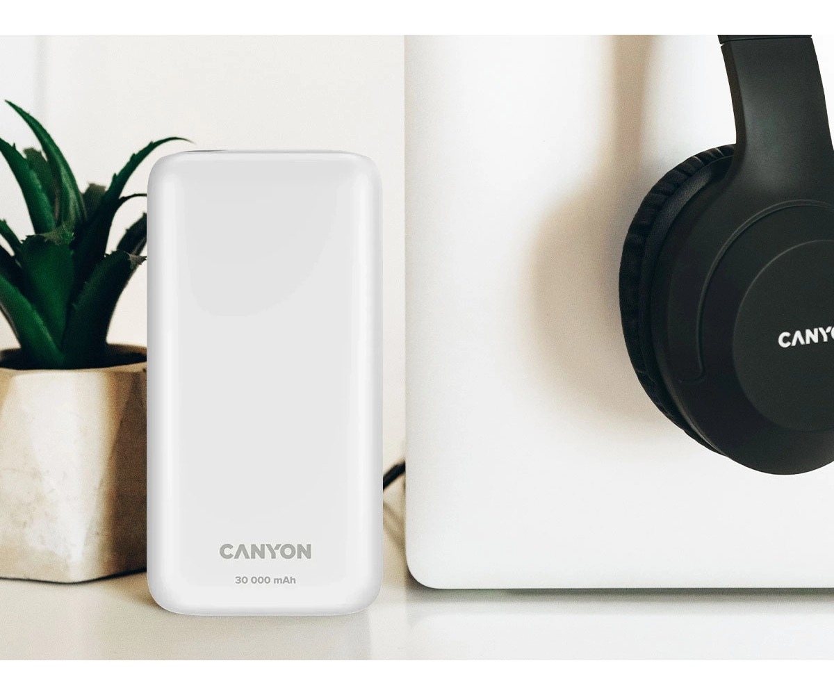 Biały powerbank Canyon (30 000 mAh) na stole, obok rośliny w doniczce i czarnych słuchawek.