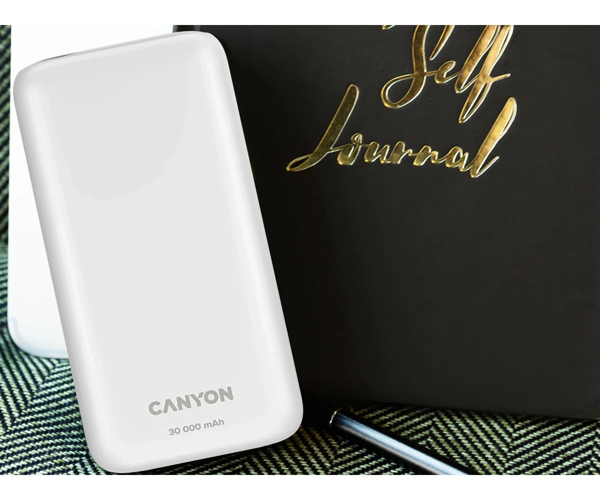 Biały powerbank Canyon o pojemności 30 000 mAh stoi obok czarnego dziennika i długopisu na wzorzystej powierzchni.
