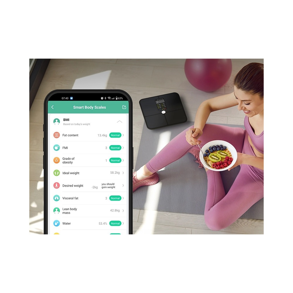 Kobieta w różowym stroju sportowym je owoce z miski, podczas gdy smartfon na pierwszym planie wyświetla dane z wagi.