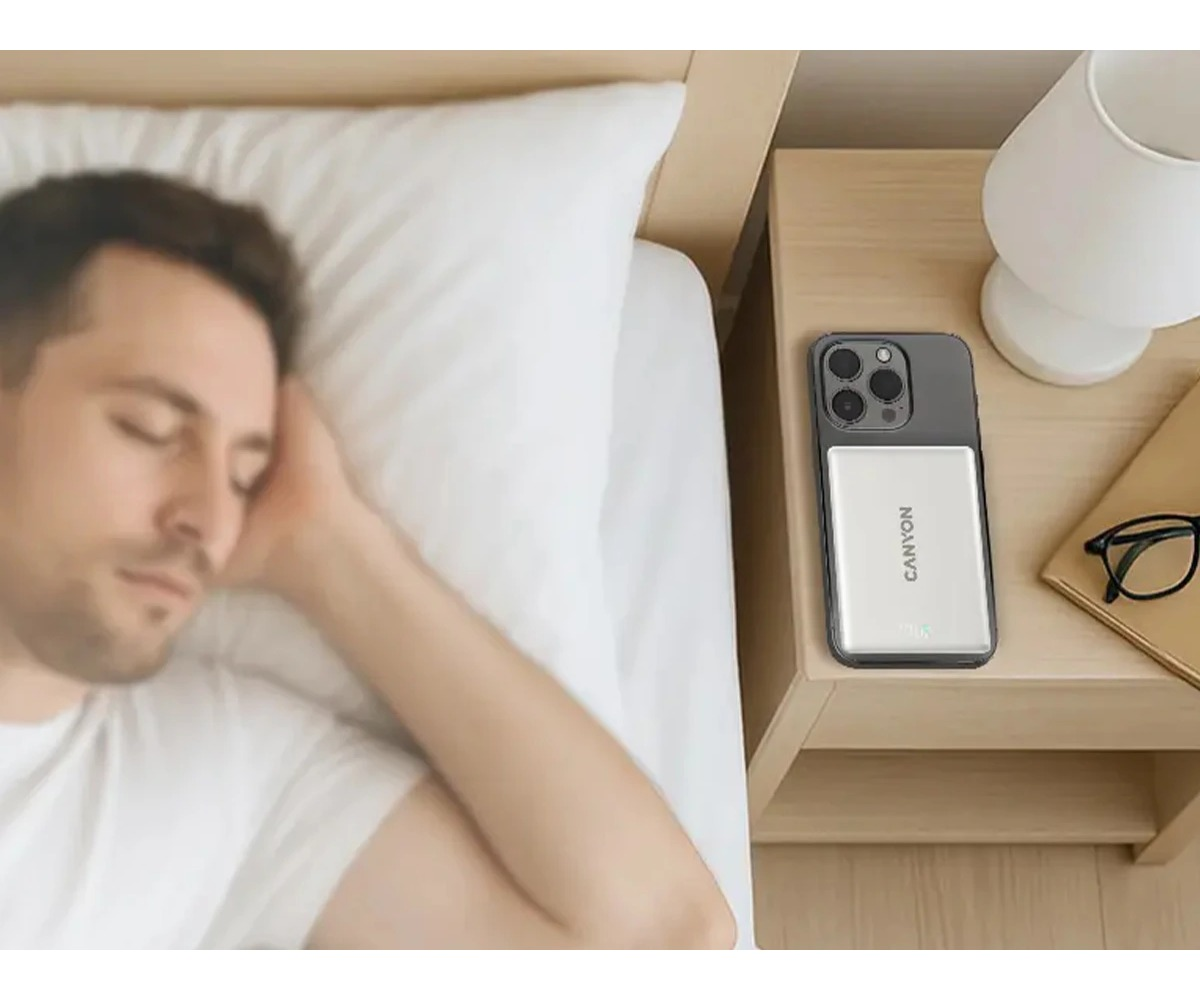 Mężczyzna śpi w łóżku, a na szafce nocnej obok niego leży smartfon z powerbankiem, lampka i okulary.