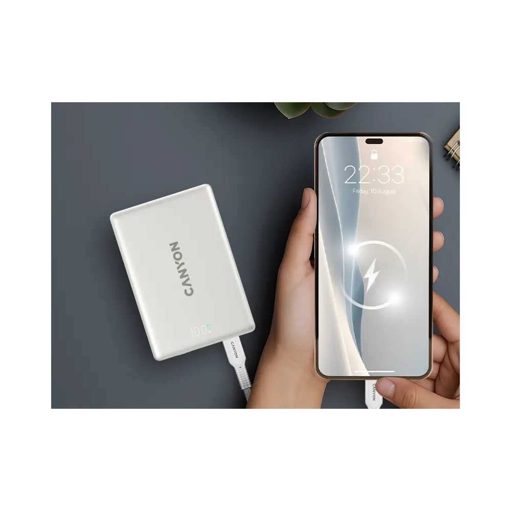 Osoba trzyma smartfon ładowany z białego powerbanku Canyon na ciemnoszarym powierzchni.