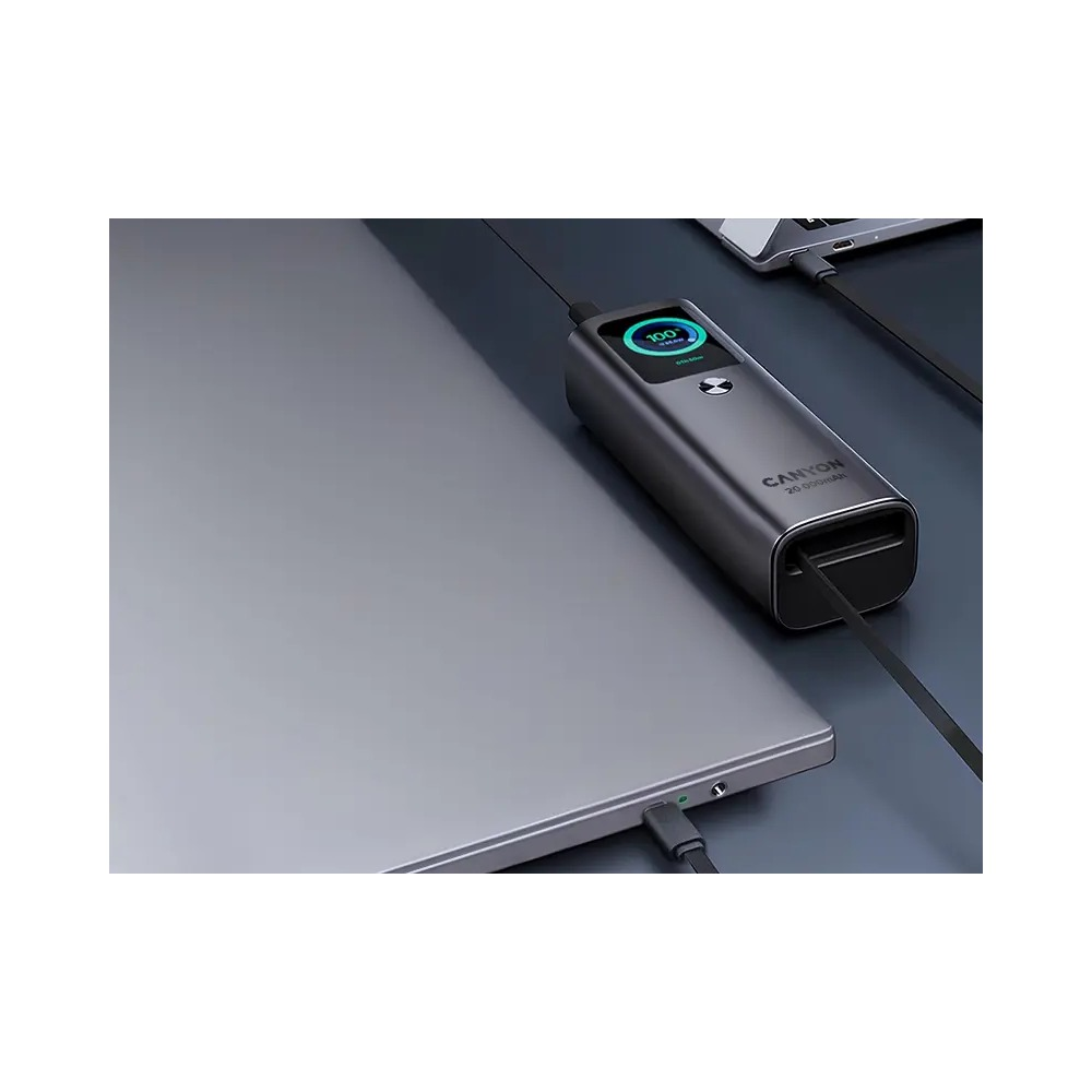 Szary powerbank Canyon z cyfrowym wyświetlaczem ładuje się na ciemnym biurku obok laptopa.