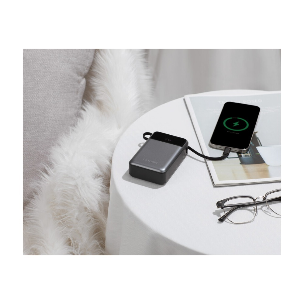 Powerbank ładuje smartfon na okrągłym stole obok okularów i poduszek.