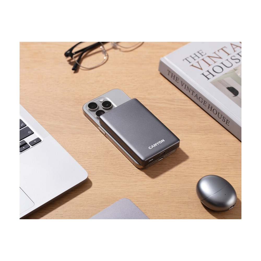 Szary powerbank Canyon jest przymocowany do smartfona na drewnianym biurku z laptopem i książką.