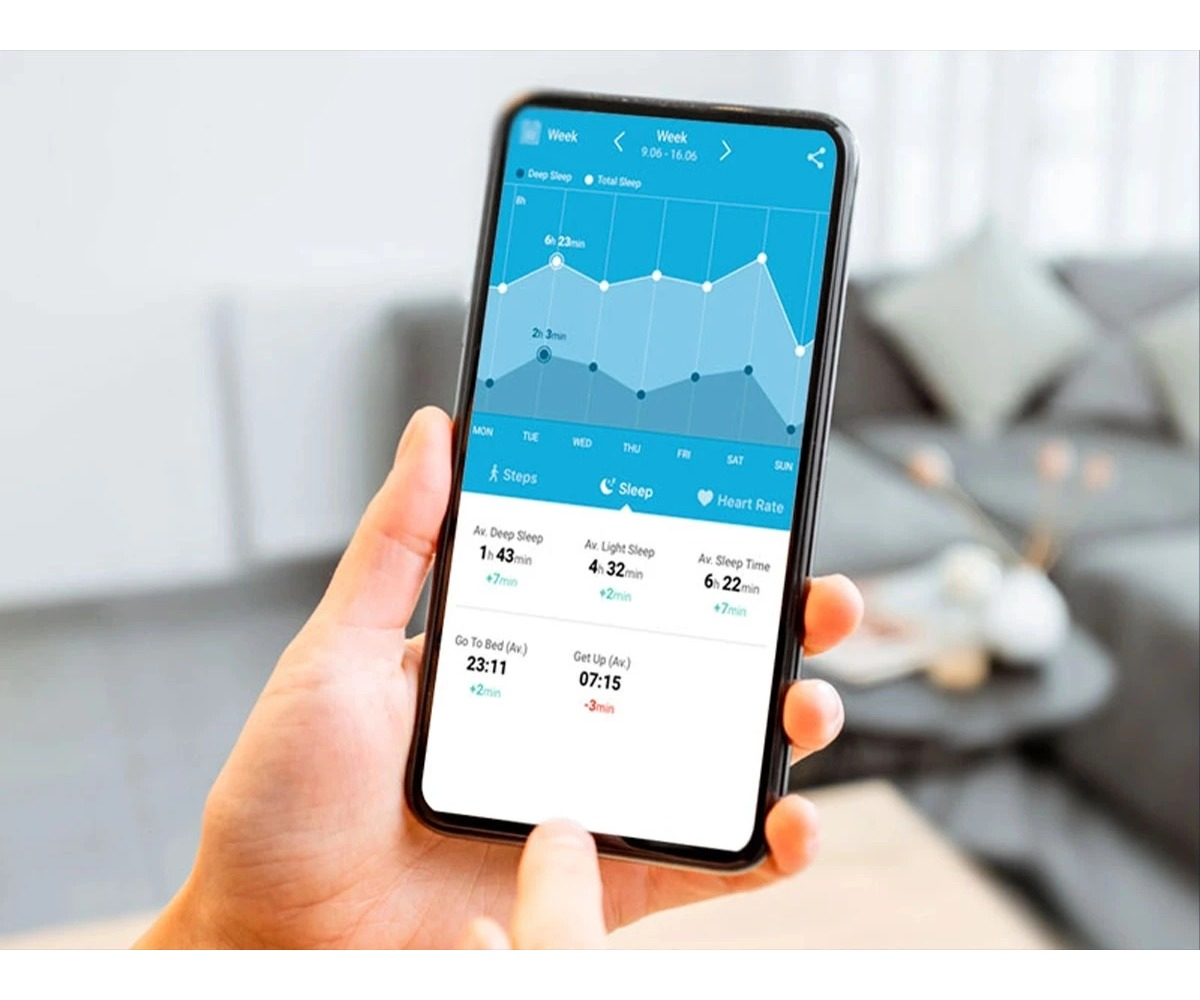 A hand holds a smartphone displaying a sleep tracking app with a blue graph screen in a living room.