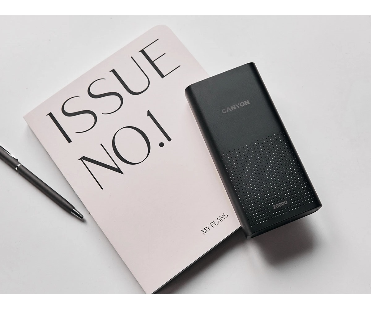 Czarny powerbank Canyon leży na różowym notesie obok długopisu na białej powierzchni.
