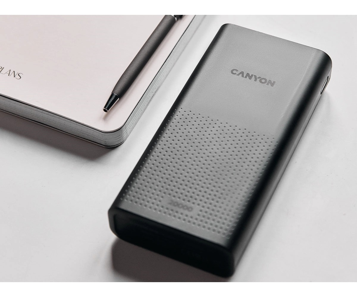 Szary powerbank Canyon leży na białym stole obok notesu i długopisu.