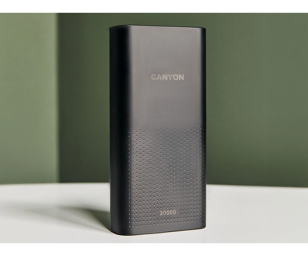 Czarny powerbank Canyon 20000 stoi na białym stole na tle zielonej ściany.