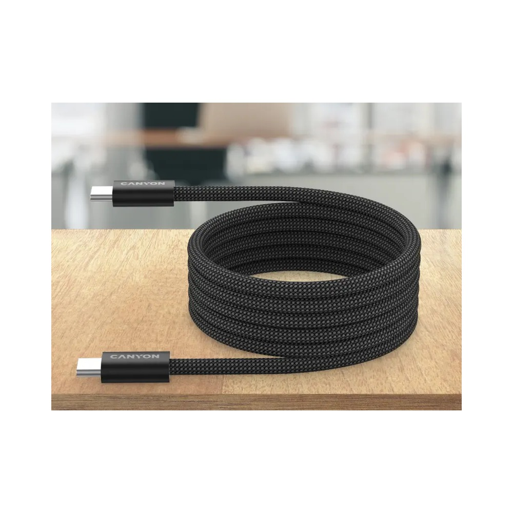 Czarny, pleciony kabel USB-C Canyon zwinięty na drewnianym blacie z rozmytym tłem.