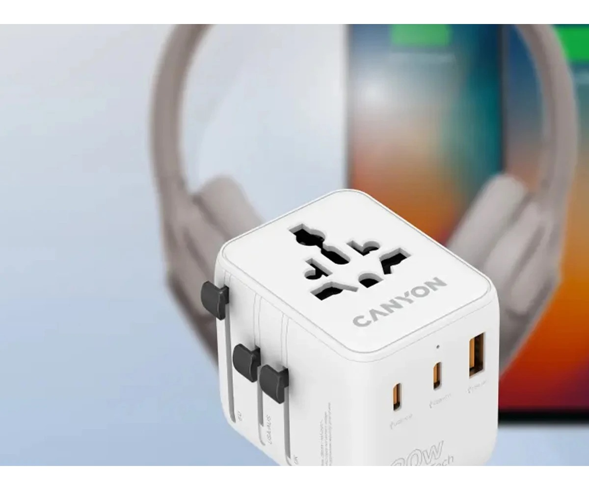Biały adapter podróżny Canyon z portami USB na jasnym tle z rozmytymi słuchawkami i telefonem.